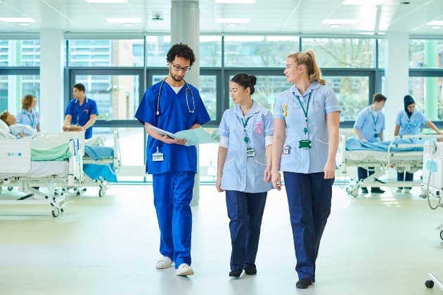 Three medical professionals walk through a bright hospital ward, discussing a patient's chart together.