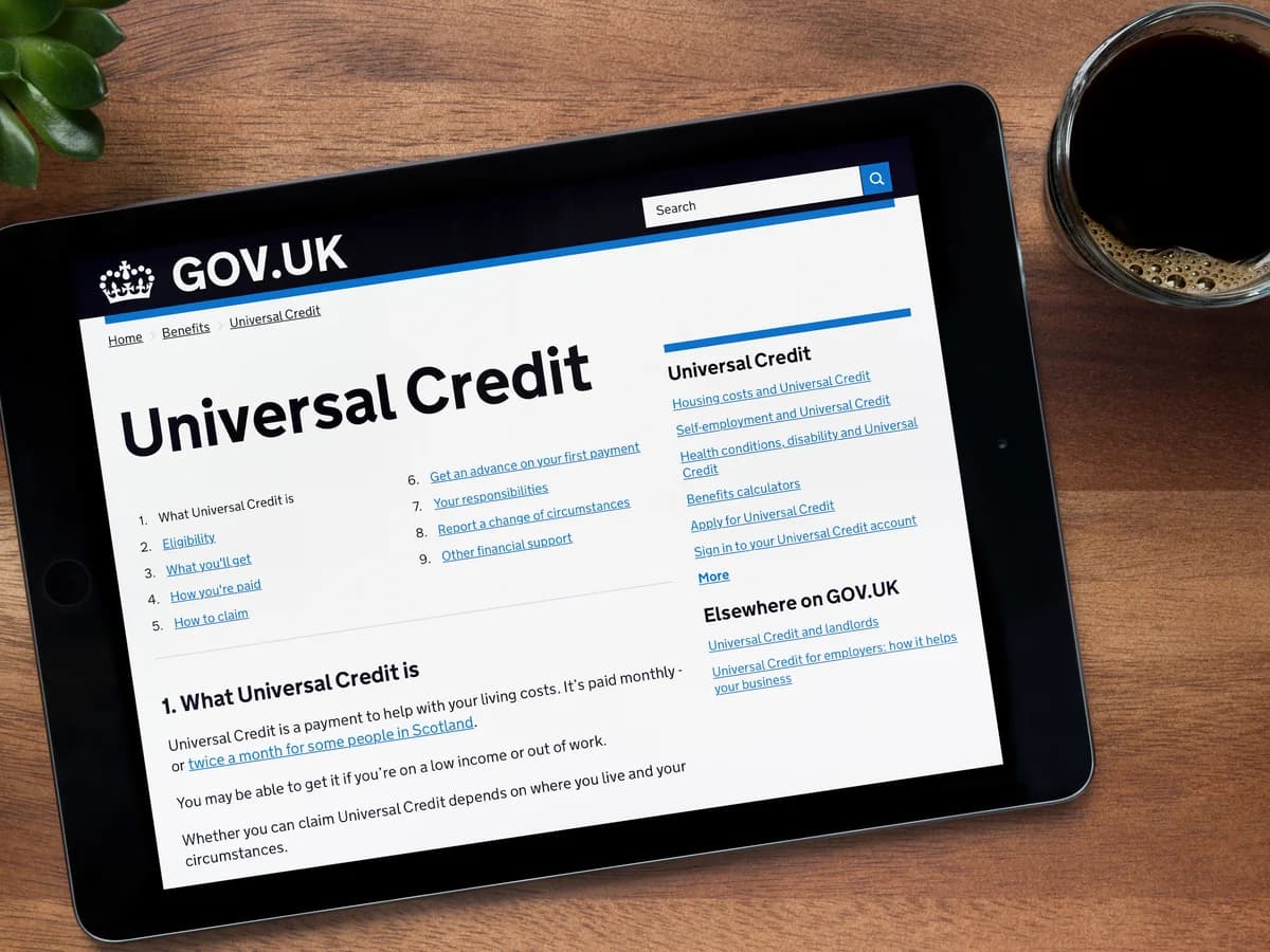Tablet showing the GOV.UK Universal Credit website on a wooden desk with coffee and plant.